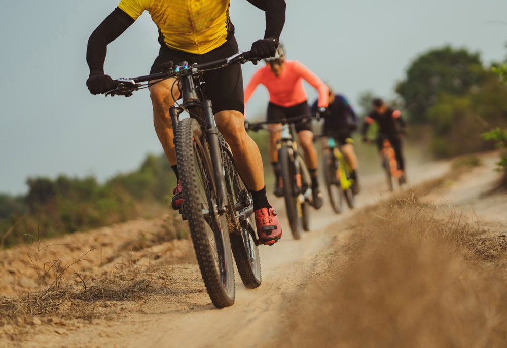 Cyclists ride mountain bikes on a dirt trail in a line.