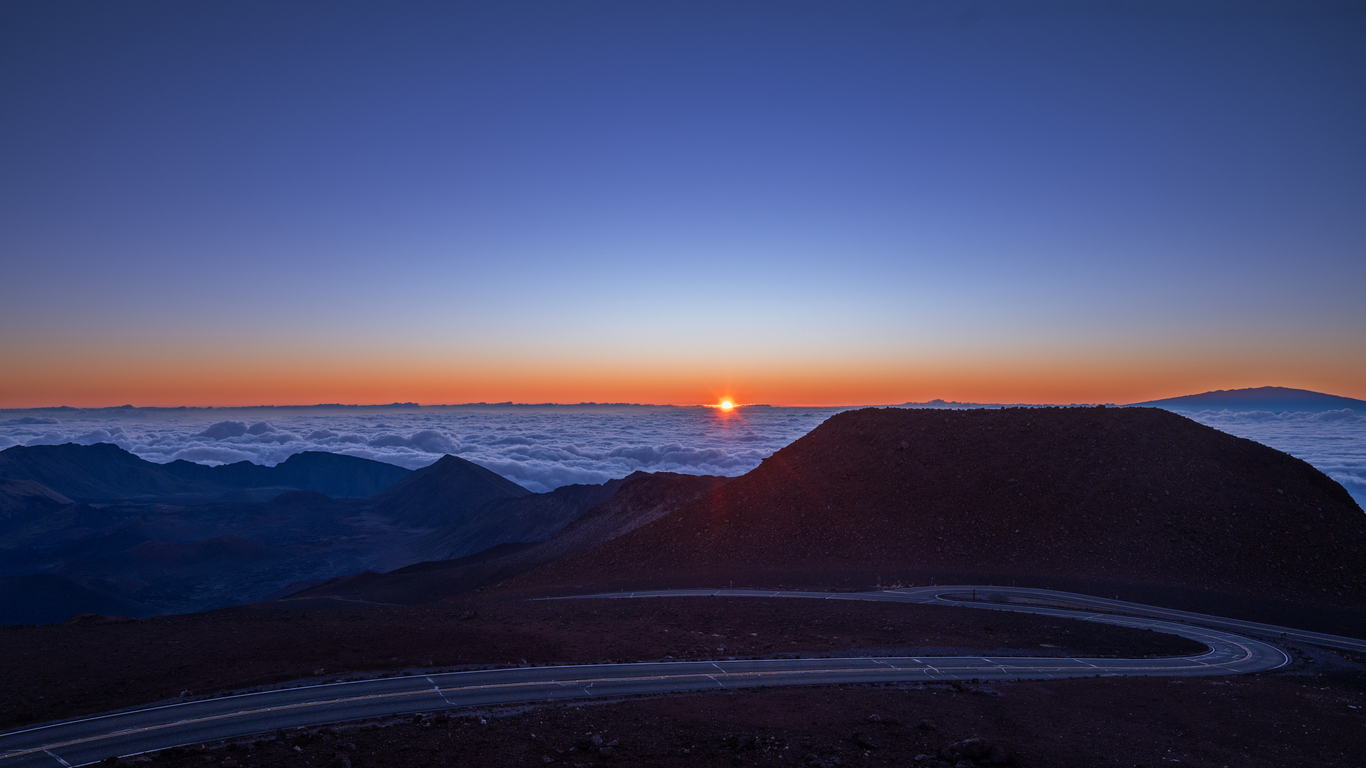 Sunrise over mountains with clouds below and a winding road in the foreground.