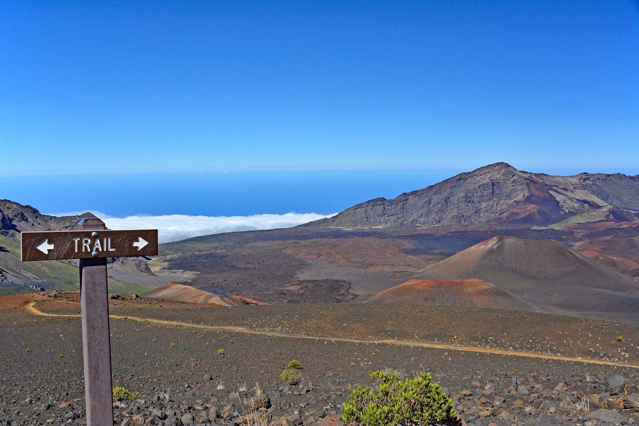 Trail sign in volcanic landscape with mountains and blue sky in background.