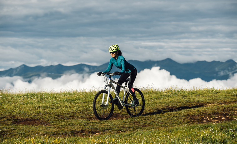 Woman biking on grassy hill with mountains and clouds in background.