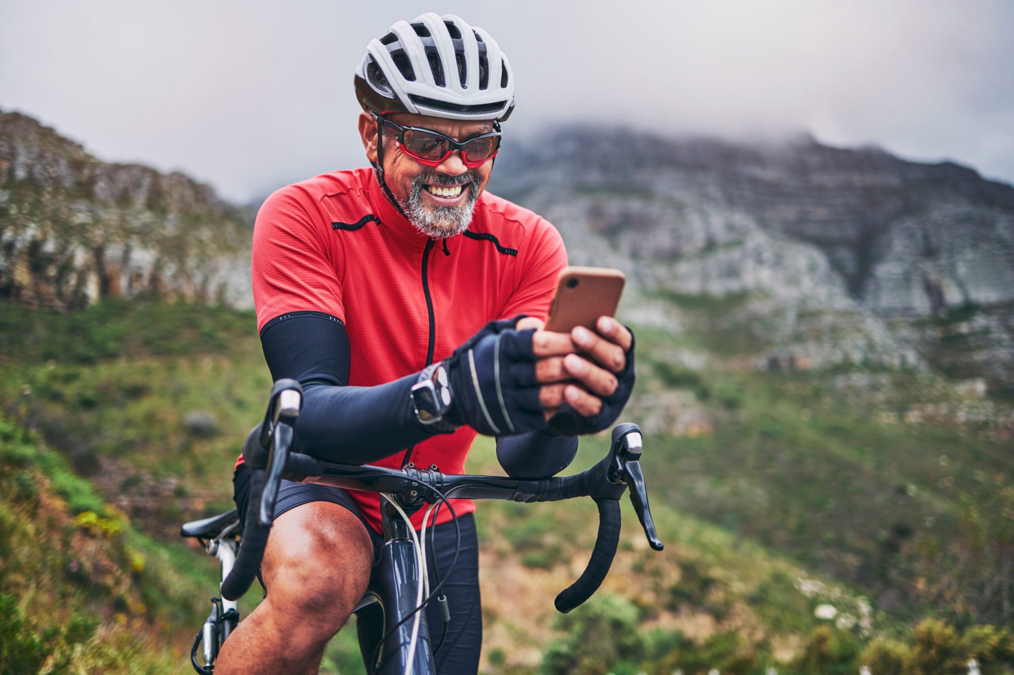 Cyclist in red jersey checks phone while smiling, mountain landscape in background.