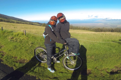 Two people in helmets on a bicycle in a grassy landscape with clear blue sky.
