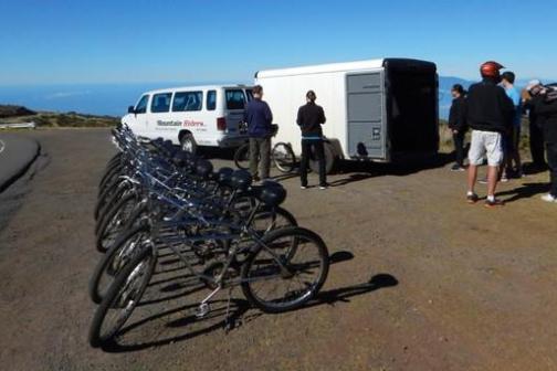 Row of bikes by a van and trailer on a scenic hilltop with people gathered nearby.