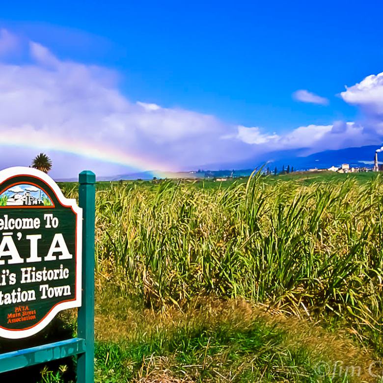 Sign for Pāʻia, Maui's Historic Plantation Town, with sugar cane field and rainbow in background.