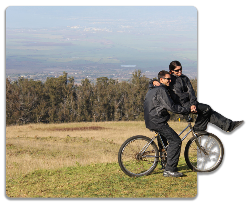 Two people on a bicycle in a grassy field with trees and a distant view.